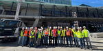 Gruppenfoto von Exkursionsteilnehmenden mit Warnwesten und Bauhelmen vor Hochhausbaustelle