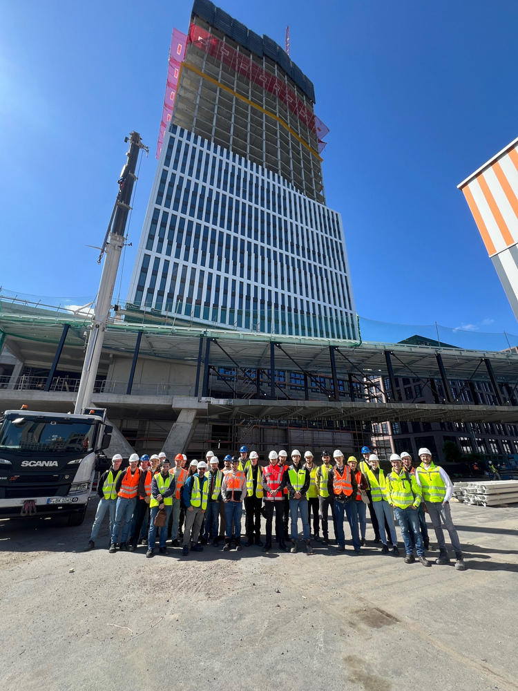 Gruppenfoto von Exkursionsteilnehmenden mit Warnwesten und Bauhelmen vor Hochhausbaustelle